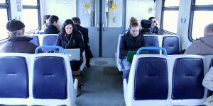 Two women work on their laptops in a train car with empty blue seats in the foreground